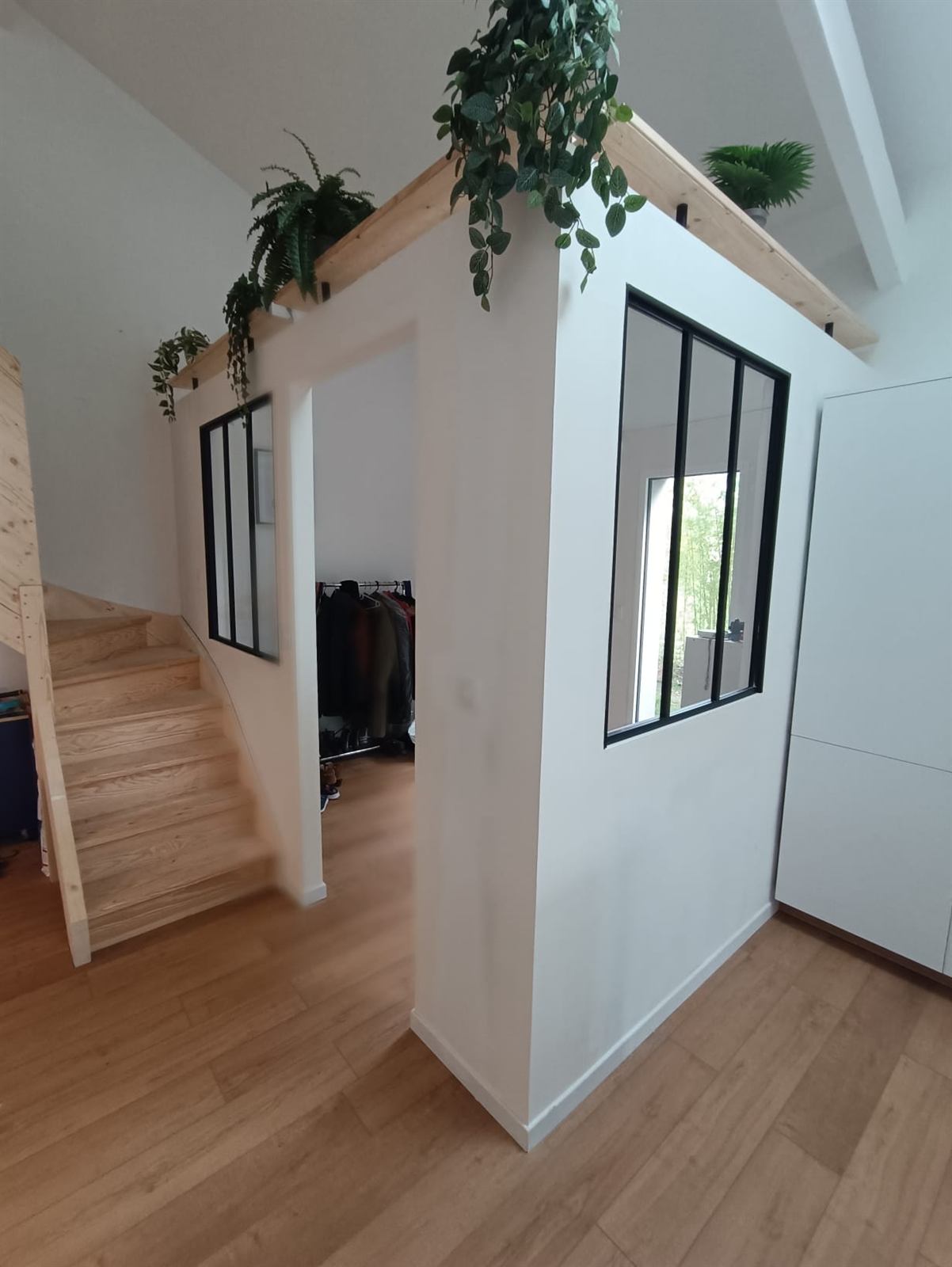 Cube en placo blanc avec verrières noires et étagère végétalisée, à côté d’un escalier en bois
